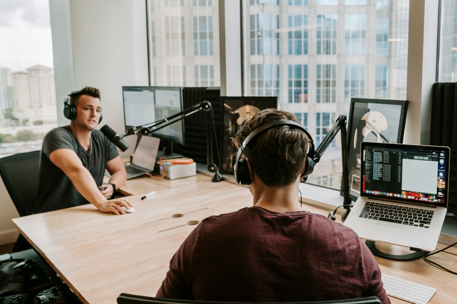Two podcast hosts in headphones talking at microphones across a desk in a bright high-rise studio.
