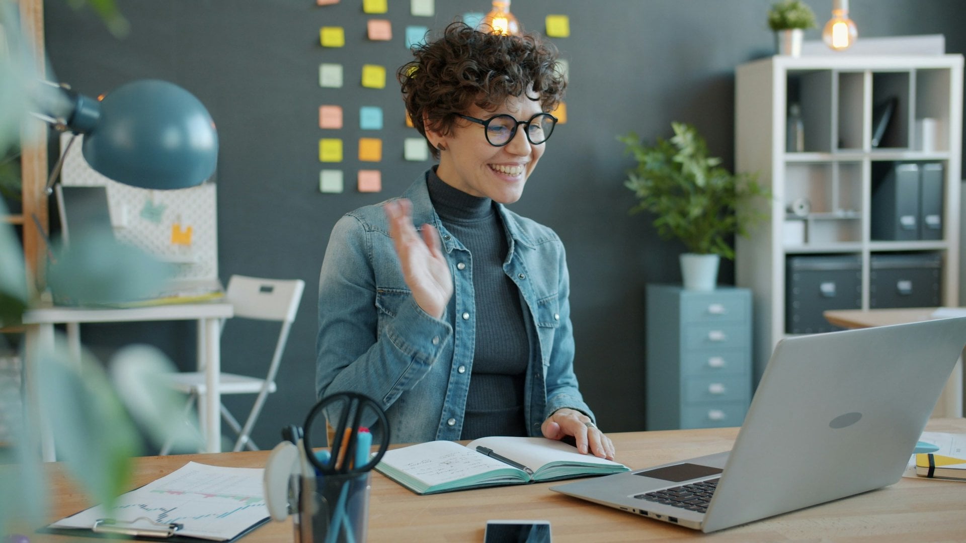 Smiling woman waving during a laptop video call at her desk in a bright home office.