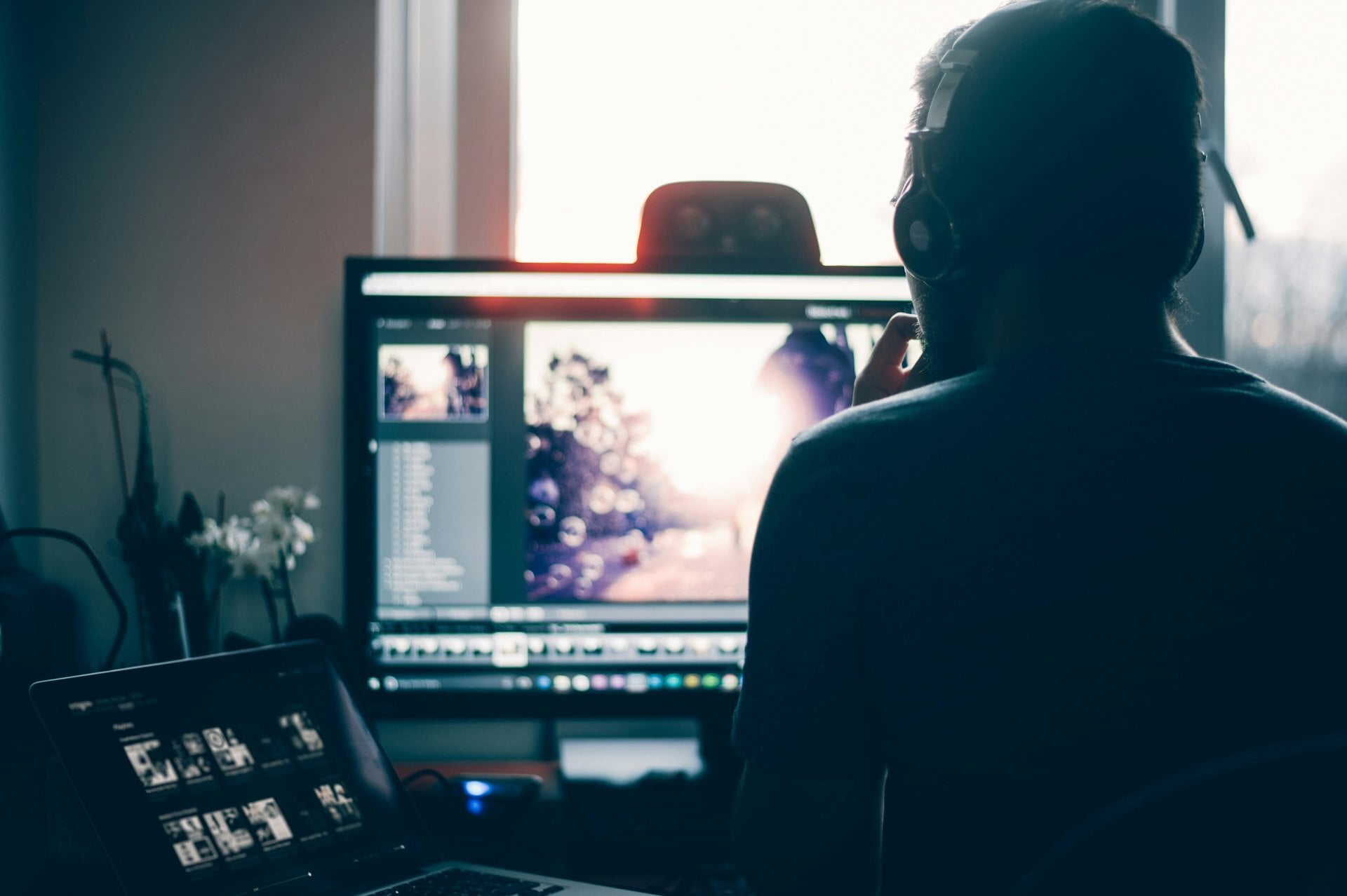 Creator in headphones editing photos on a desktop monitor beside a laptop in a softly lit home studio.